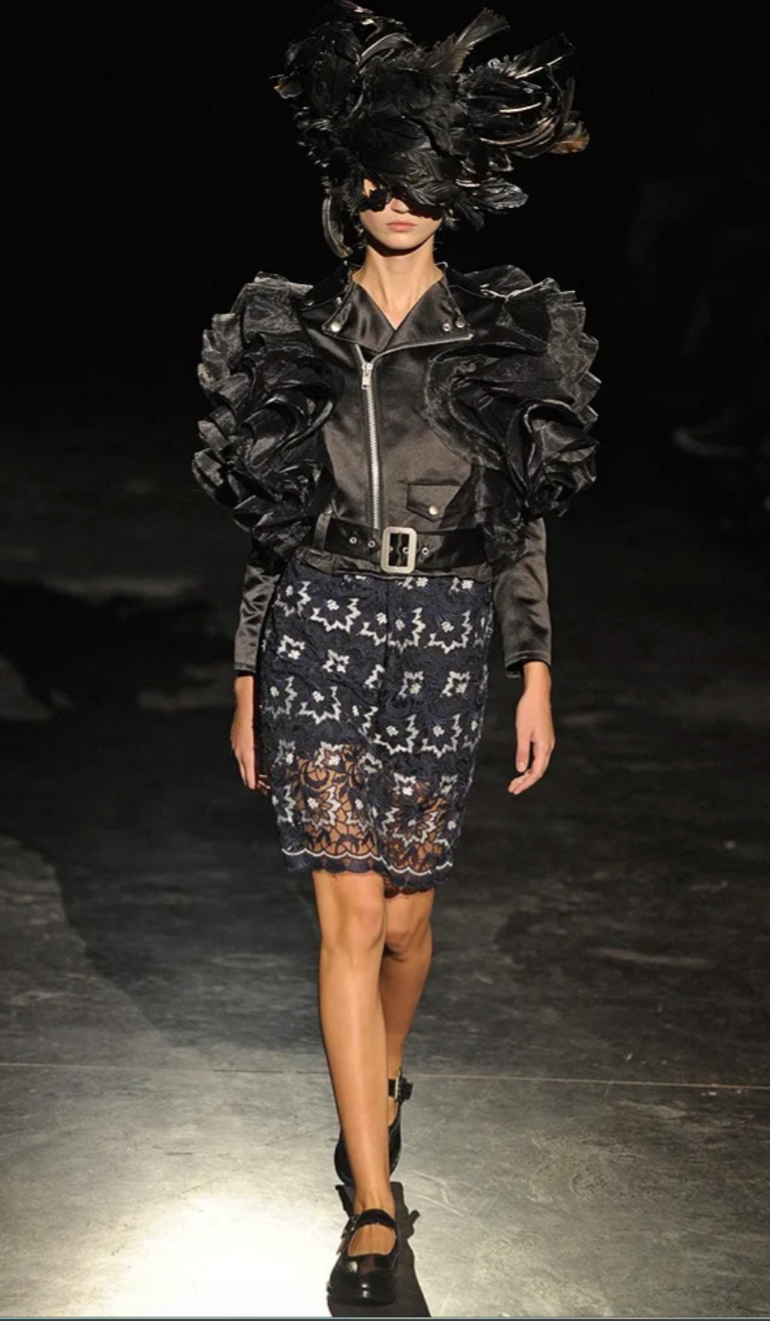 Model walking on a runway wearing a black ruffled jacket and patterned skirt.